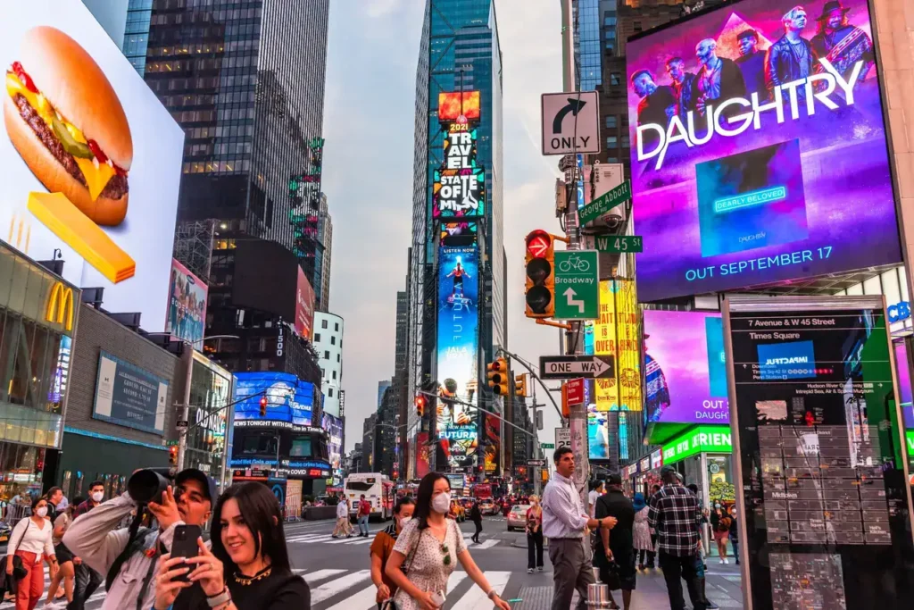 Crowd in Times Square with vibrant digital billboards showcasing advertisements, including a hamburger and a Daughtry concert promotion, highlighting urban advertising and guerrilla marketing strategies.