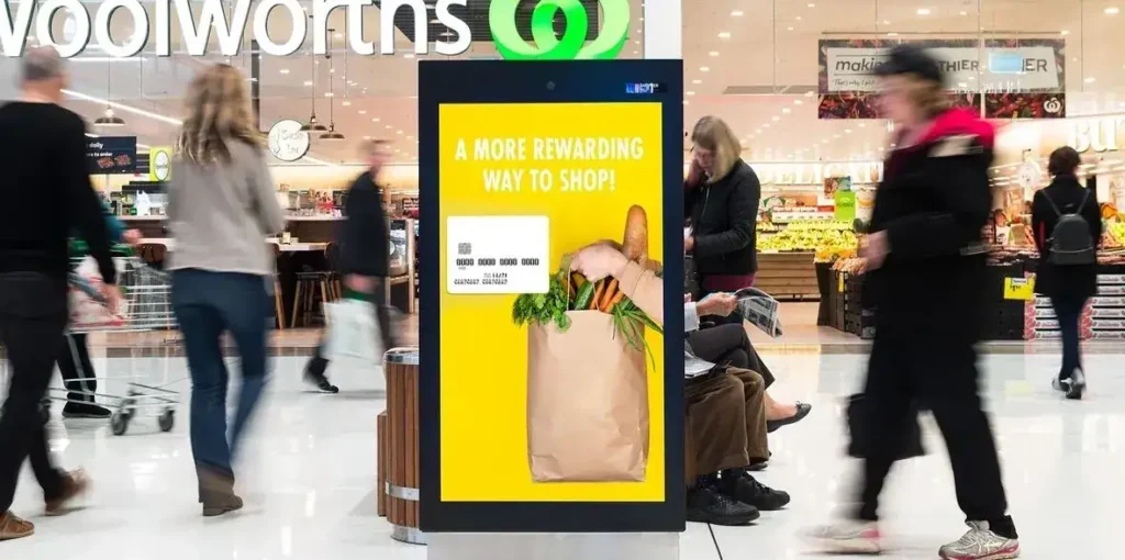 Digital advertising display promoting shopping rewards in a busy retail environment, with shoppers and grocery items visible in the background.