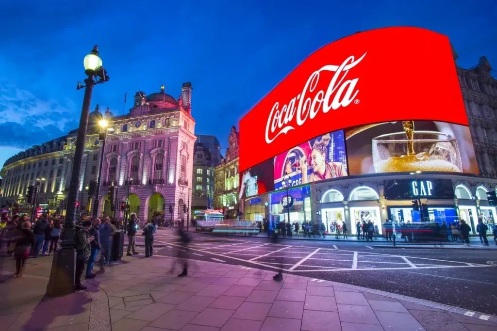 Coca-Cola advertisement on the iconic Piccadilly Lights in London, showcasing vibrant colors and bustling atmosphere, surrounded by historic architecture and pedestrians.