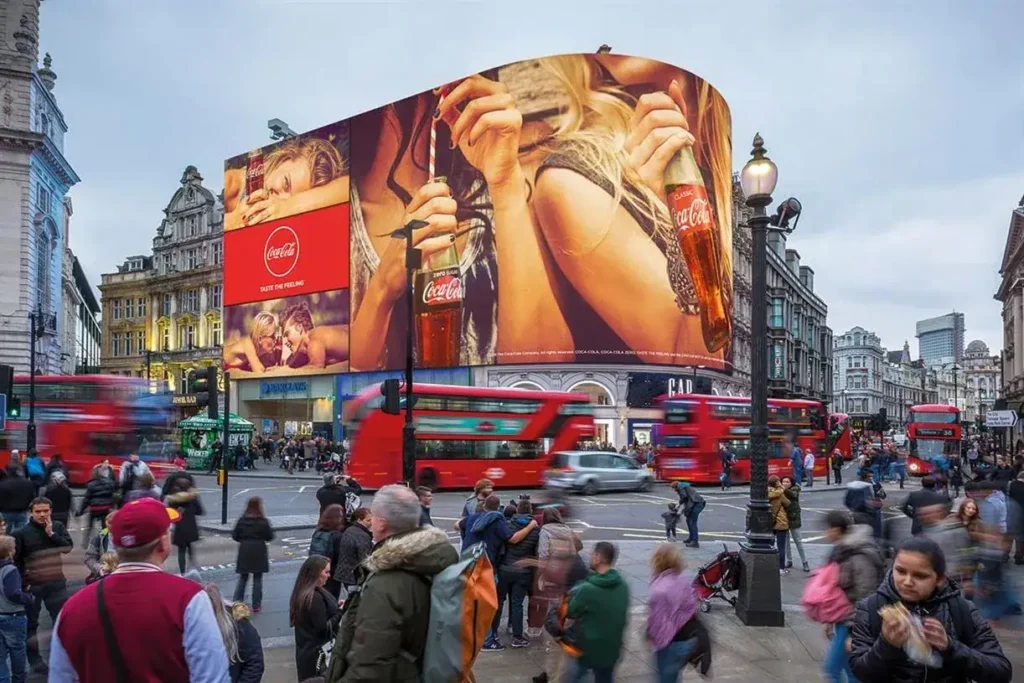 Coca-Cola advertisement on a large 3D billboard in a busy urban setting, featuring people holding bottles, with red double-decker buses and pedestrians in the foreground, showcasing international advertising in a vibrant city.
