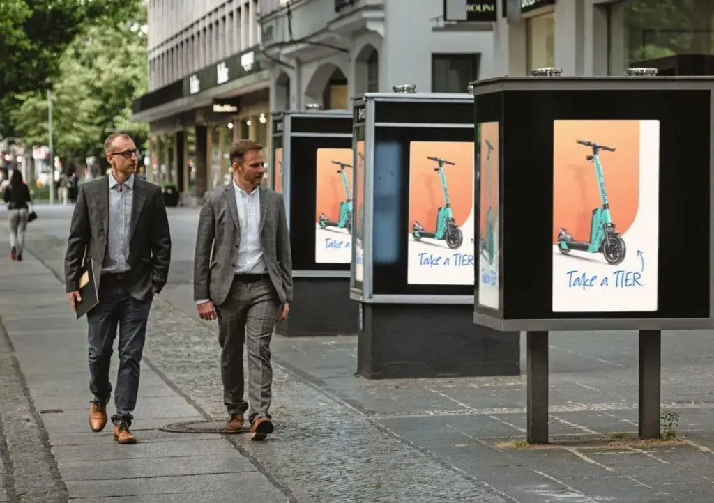 Two business professionals walking on a city street, passing advertising displays featuring electric scooters with the slogan "Take a TIER," highlighting urban mobility solutions in a culturally diverse environment.