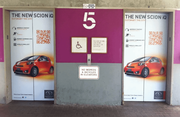Elevator doors wrapped with vibrant advertising for the new Scion iQ, featuring bold graphics, QR code, and marketing text highlighting its features, promoting brand visibility in a high-traffic area.