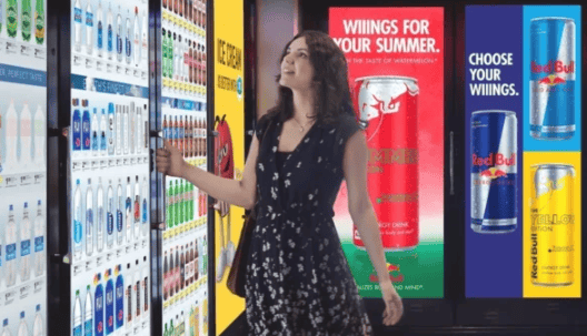 Woman selecting beverages from a vending machine, surrounded by colorful advertising for Red Bull and other drinks, illustrating retail advertising strategies.