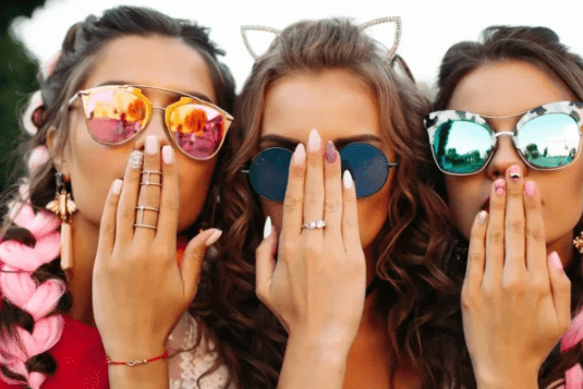 Three young women wearing stylish sunglasses, playfully covering their mouths with hands, showcasing a trendy and vibrant social atmosphere.