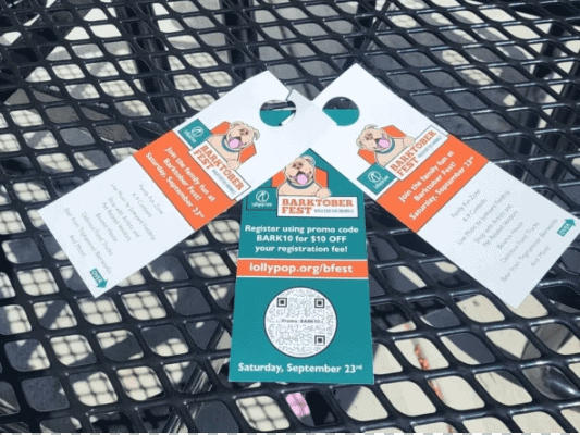 Three colorful door hangers for Barktober Fest, featuring a dog graphic, event details, and a QR code for registration, placed on a black outdoor table.