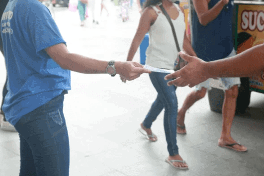 Person in blue shirt handing out a flyer to another individual in a busy urban setting, illustrating the tangible engagement of flyer distribution in marketing.