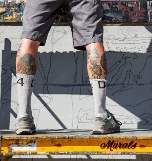 Person standing on a ladder wearing shorts and socks, showcasing detailed leg tattoos, with a mural backdrop, emphasizing artistic expression in mural advertising.