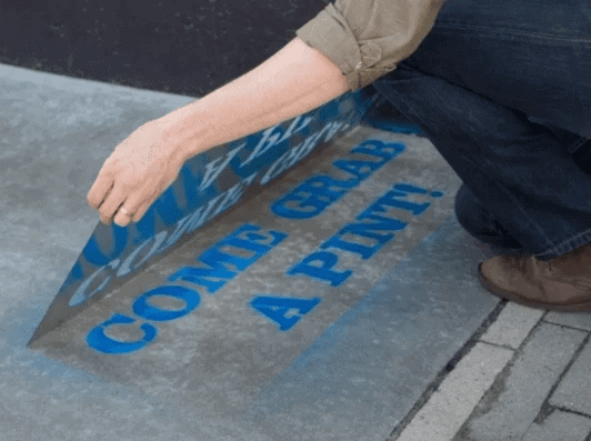 Person applying sidewalk stencil with the text "COME GRAB A PINT!" for guerrilla marketing campaign, showcasing creative advertising technique.