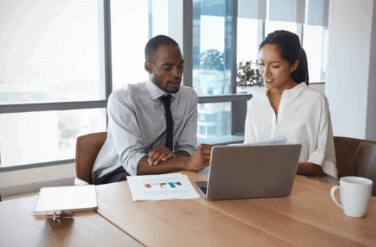 Two professionals collaborating at a table with a laptop and documents, discussing marketing strategies in a modern office setting.