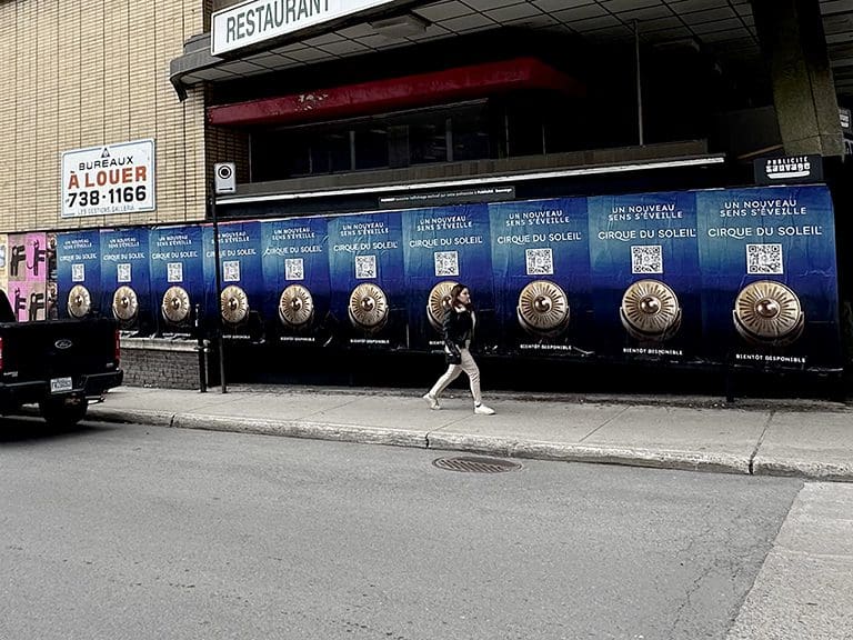 Billboard advertising for Cirque du Soleil featuring multiple large posters with a blue background and a central circular design, showcasing the phrase "UN NOUVEAU S'EST R&Eacute;VEILL&Eacute;" and a QR code, with a pedestrian walking past on a city sidewalk.