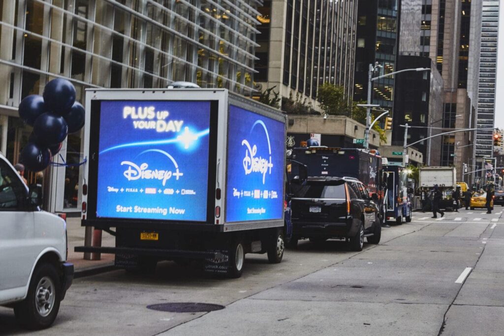 Disney+ LED billboard truck advertising with promotional text "PLUS UP YOUR DAY" and "Start Streaming Now" on city street.