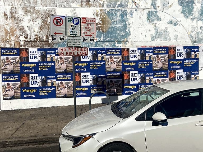 White car parked near a wall covered with Wrangler advertisements promoting "Get Up" and "Longhorn Legendary," alongside parking signs indicating restrictions.