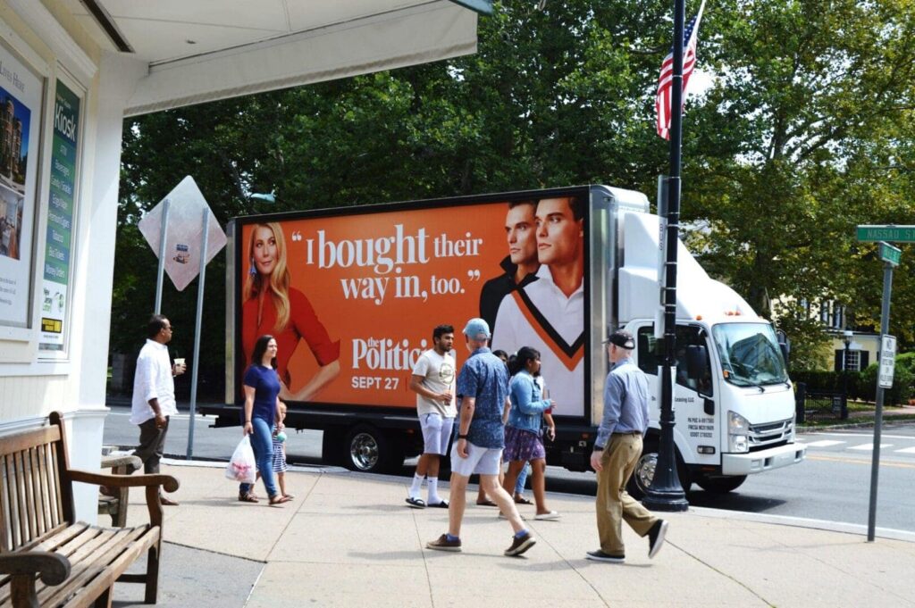 Mobile billboard truck displaying political advertisement featuring three individuals with the text "I bought their way in, too" and the date "SEPT 27," surrounded by pedestrians in an urban setting.