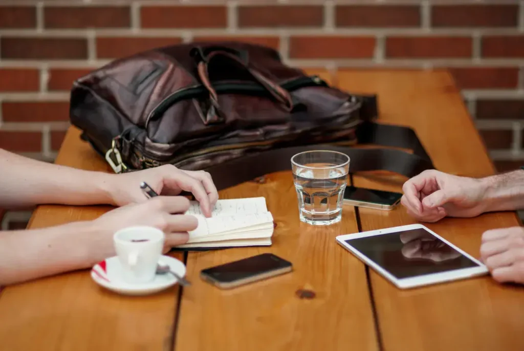 Hands writing notes and discussing strategies over a table with a laptop, smartphone, coffee cup, and water glass, reflecting collaborative digital advertising planning.