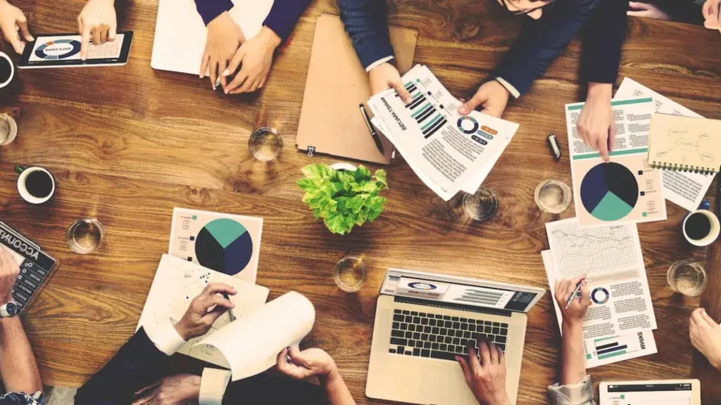 Group of professionals collaborating at a wooden table with marketing reports, charts, and a laptop, illustrating media planning and advertising strategies.