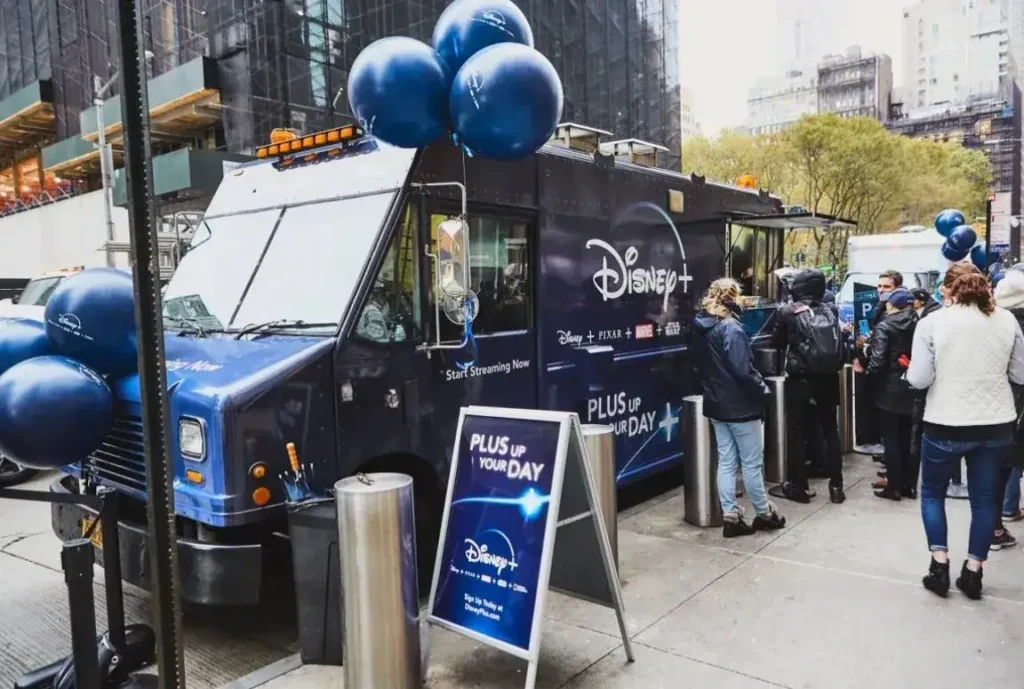 Disney+ branded food truck with promotional balloons and signage, featuring the slogan "Plus Up Your Day," attracting a crowd in an urban setting.