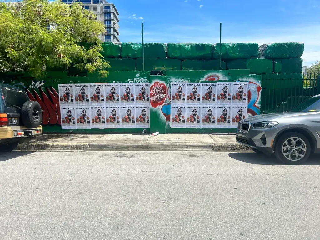 Urban wall covered with promotional posters for guerrilla marketing, surrounded by vehicles and green foliage, emphasizing street-level advertising strategies.