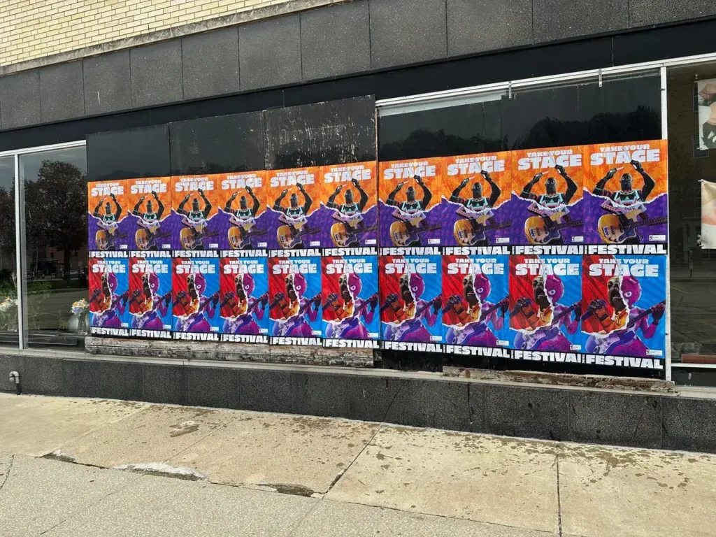 Colorful promotional posters for a festival displayed on a storefront window, featuring bold graphics and repeated text emphasizing "STAGE" and "FESTIVAL."