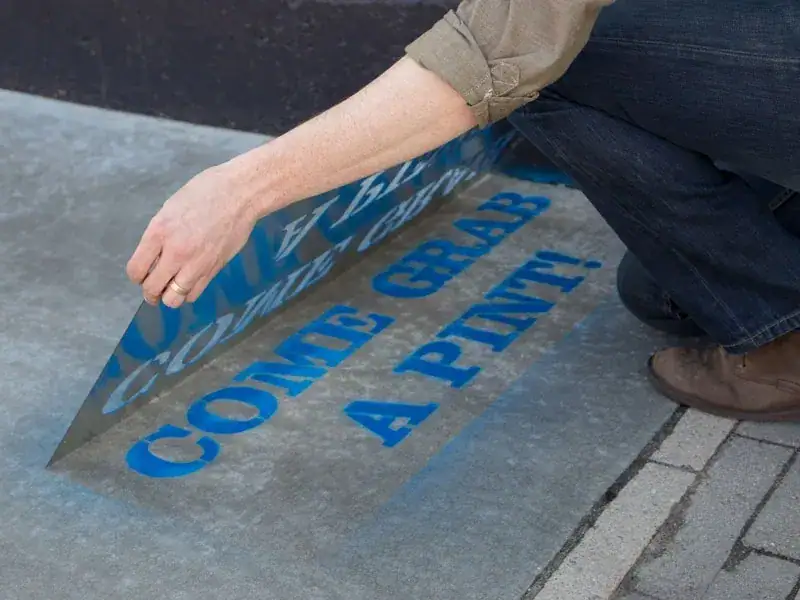 Person applying sidewalk stencil with the text "COME GRAB A PINT!" for local marketing promotion, relevant to guerrilla marketing strategies.