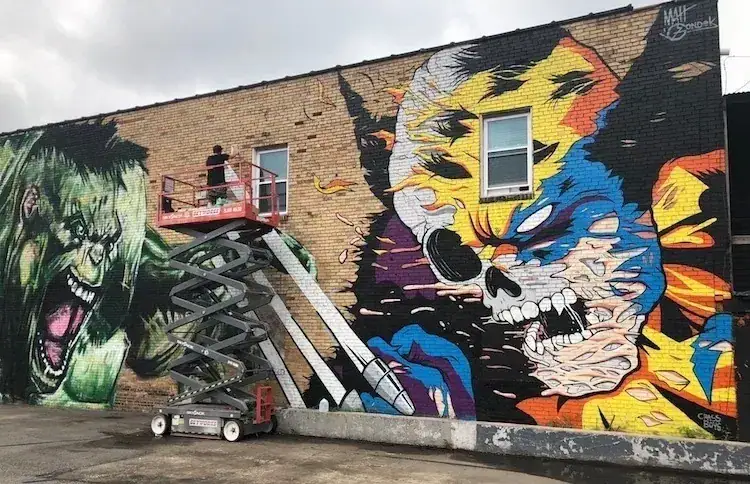 Mural artist painting vibrant graffiti featuring a skull and a green monster on a brick wall, showcasing creative mural advertising techniques.