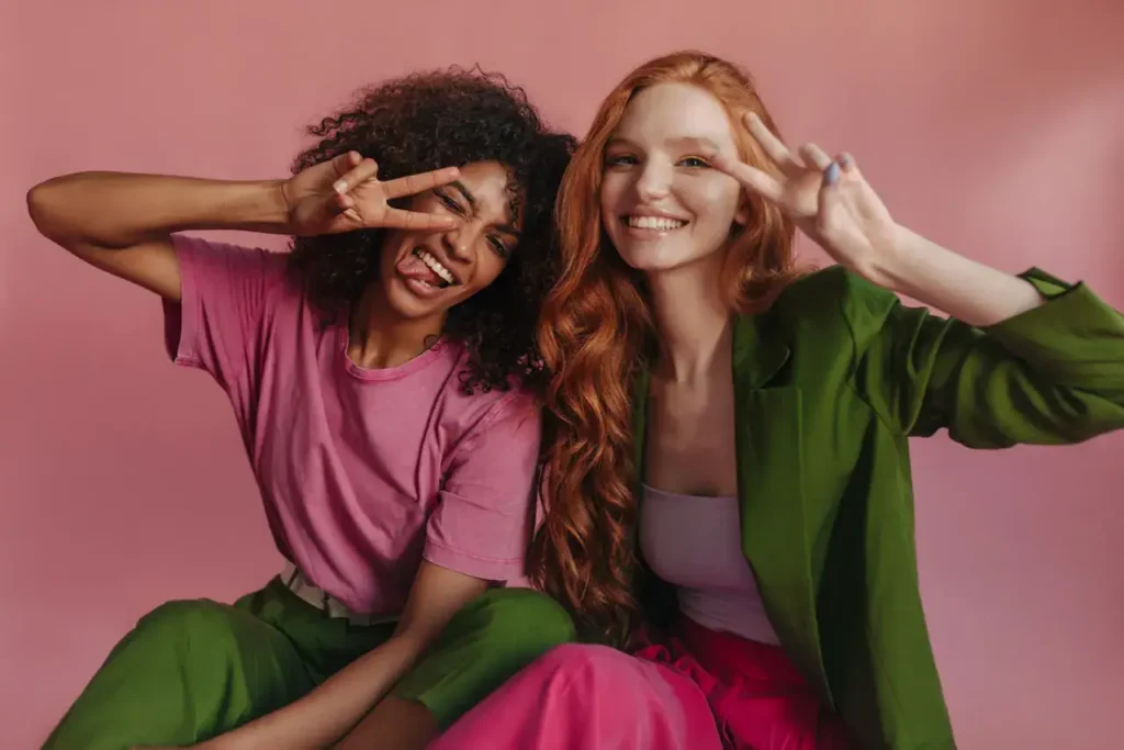 Two women smiling and posing with peace signs, wearing colorful outfits against a pink background, representing a vibrant and engaging brand ambassador image.