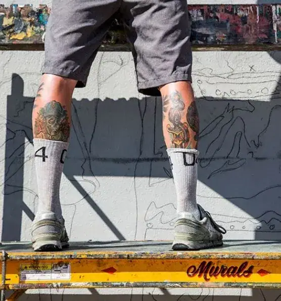 Person standing on a ladder, wearing shorts and socks with the number "40," showcasing colorful tattoos on legs, preparing to create mural art in an urban setting.