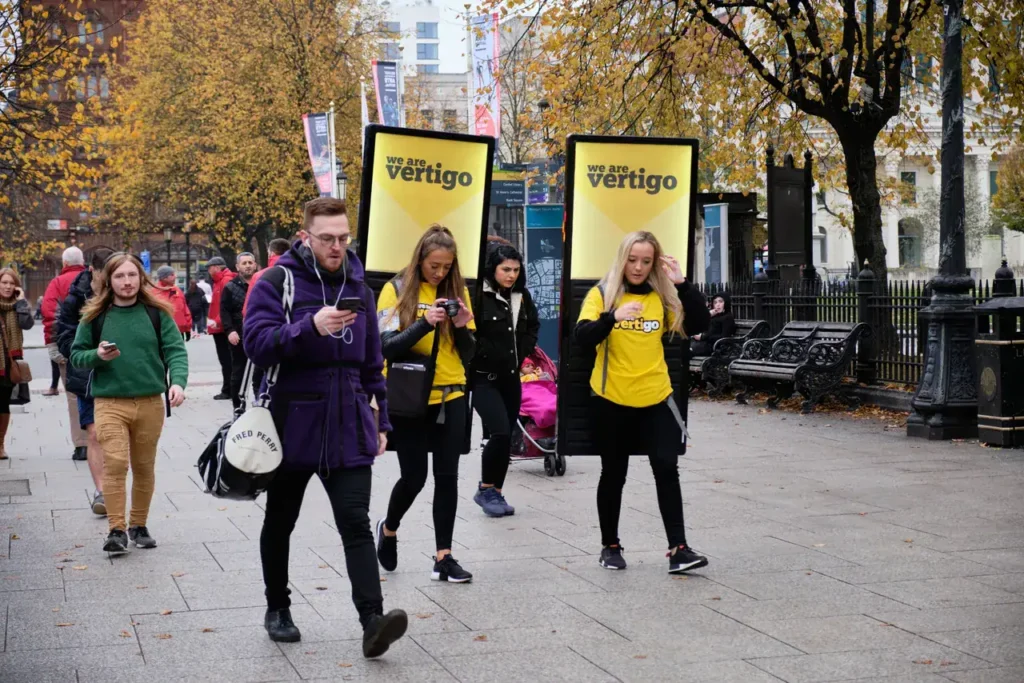 People walking in a city street, two women in yellow shirts promoting "we are vertigo" alongside a man in a purple jacket, with autumn foliage and urban scenery in the background, illustrating guerrilla marketing in action.