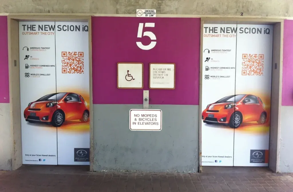 Elevator doors featuring advertising for the new Scion iQ, highlighting its compact size with phrases like "America's tiniest" and "world's smallest," alongside QR codes and branding elements.