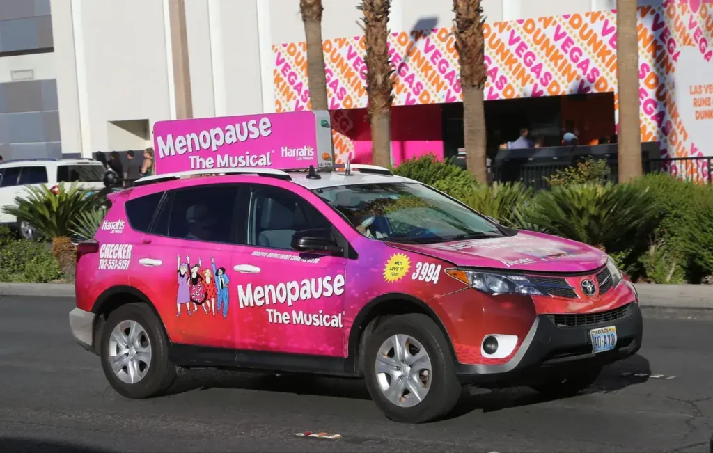Taxi advertising vehicle promoting "Menopause: The Musical" with vibrant pink graphics, parked in a lively urban setting.