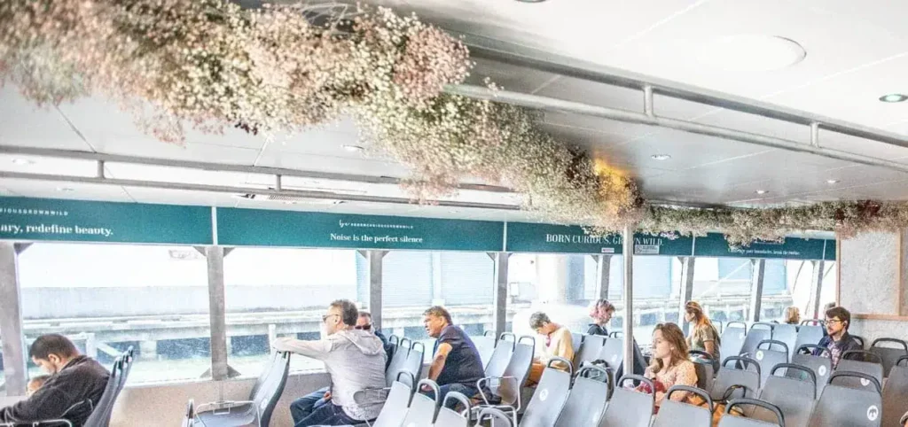 Interior of a ferry featuring decorative floral arrangements above seating, with passengers engaged in various activities, reflecting innovative transit advertising strategies.