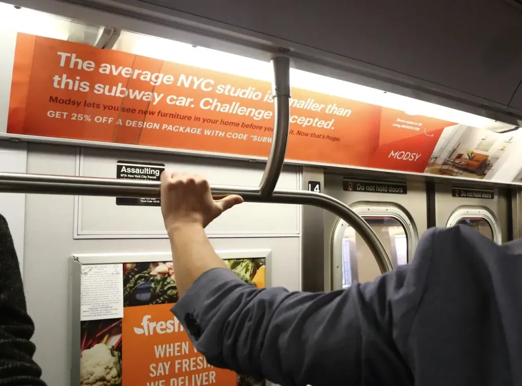 Person holding subway pole in NYC subway car, featuring advertisement for Modsy stating "The average NYC studio is smaller than this subway car. Challenge accepted." and promoting a design package discount.