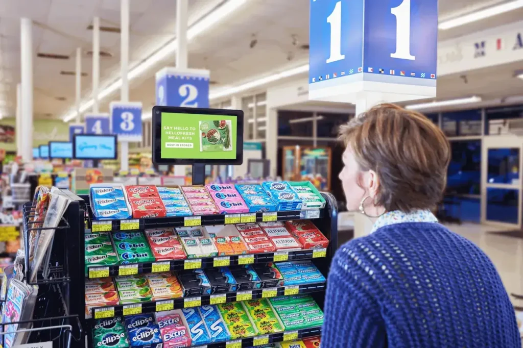 Woman viewing digital advertisement near gum display in grocery store, showcasing innovative retail advertising strategies.