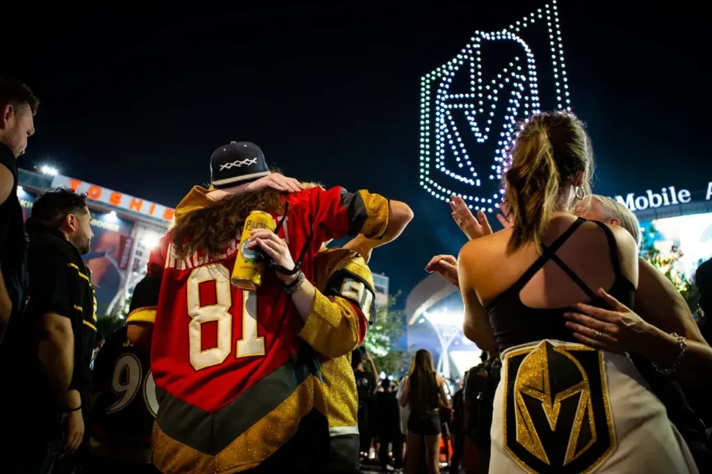 Fans celebrating at a night event, wearing Vegas Golden Knights jerseys, with a large illuminated team logo in the background.