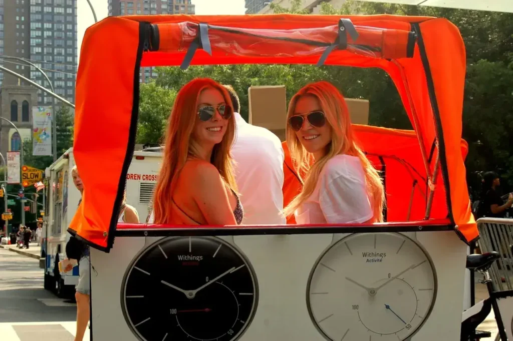 Two women smiling while seated in a pedicab with a bright orange canopy, featuring visible clocks on the front, in an urban setting.