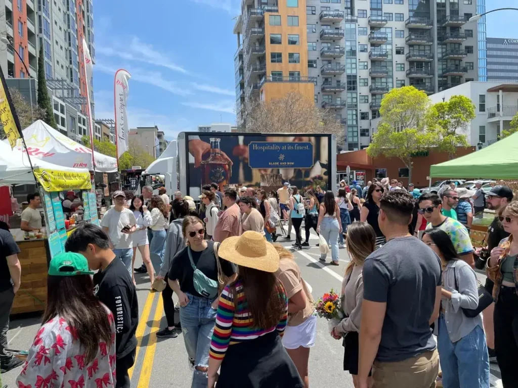 Crowd enjoying a vibrant outdoor event, with food stalls and a digital billboard displaying "Hospitality at its finest" in a bustling urban setting.