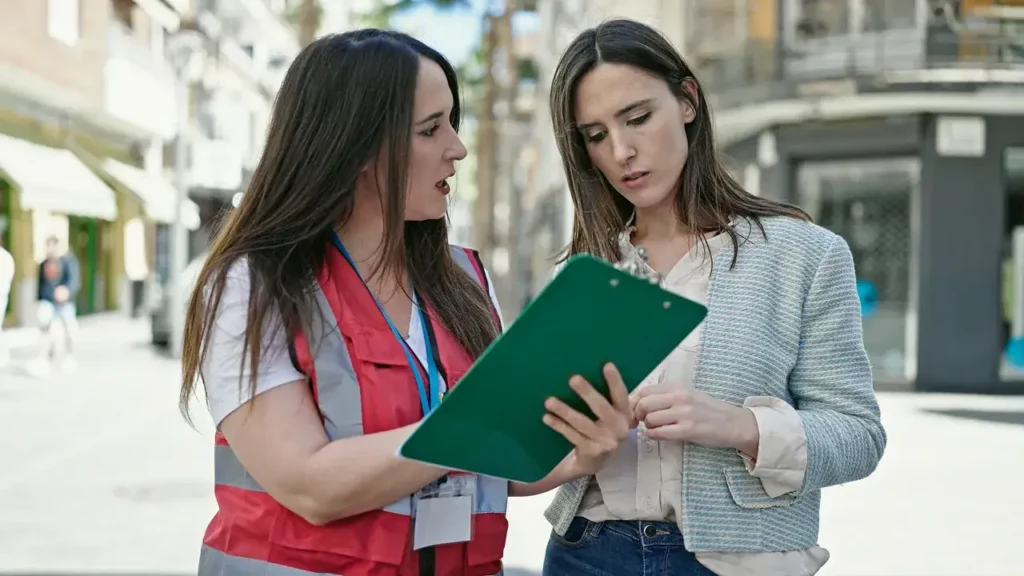 Two women discussing event staffing details, one holding a clipboard and wearing a red vest, in an urban setting.