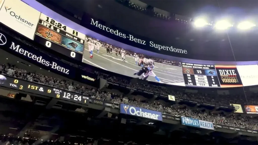 Mercedes-Benz Superdome interior during a football game, featuring large digital scoreboard displaying game stats and advertisements, with fans in attendance.