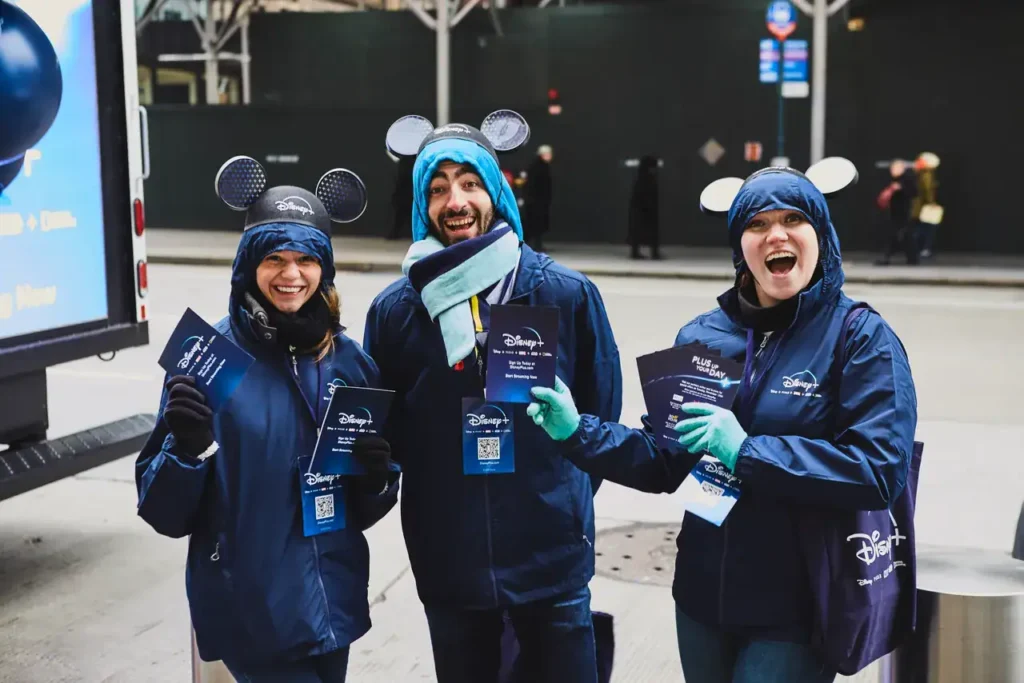 Three smiling individuals in Disney-branded jackets and Mickey Mouse ears, holding promotional materials, engaged in a guerrilla marketing campaign on a city street.