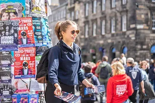 Young woman distributing flyers in a busy urban setting, surrounded by colorful posters and a crowd, highlighting effective flyer distribution strategies for marketing.