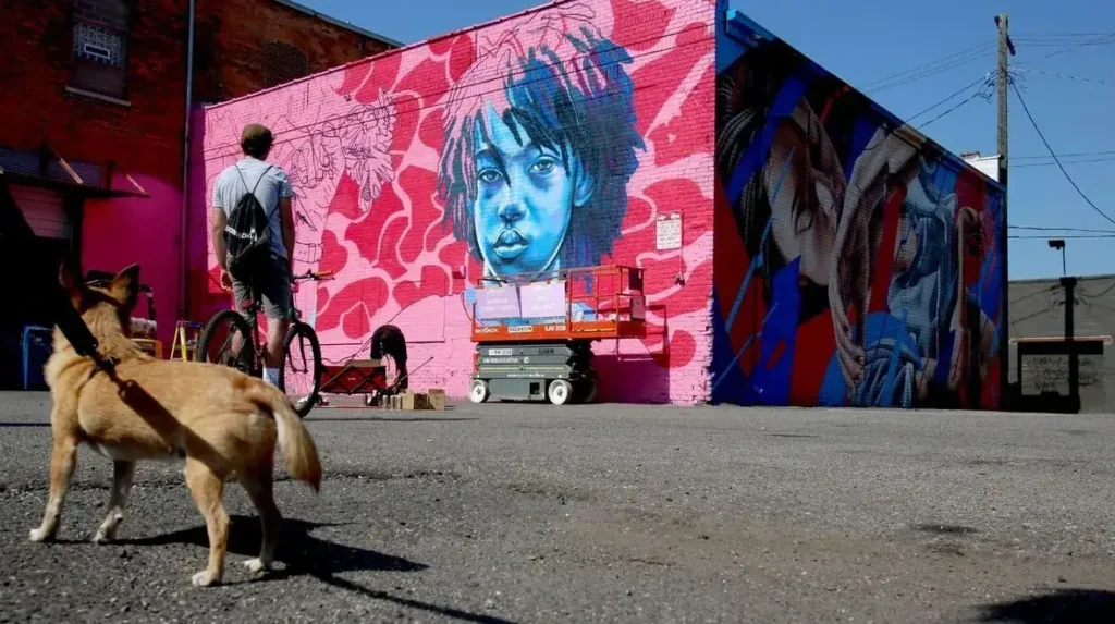 Person walking a dog in front of a vibrant mural featuring a large blue-faced portrait and colorful abstract designs, illustrating urban art and guerrilla marketing potential.