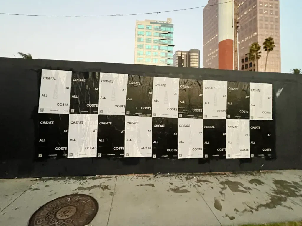 Posters displaying the phrase "CREATE AT ALL COSTS" on a black wall, part of urban advertising strategy by American Guerrilla Marketing.