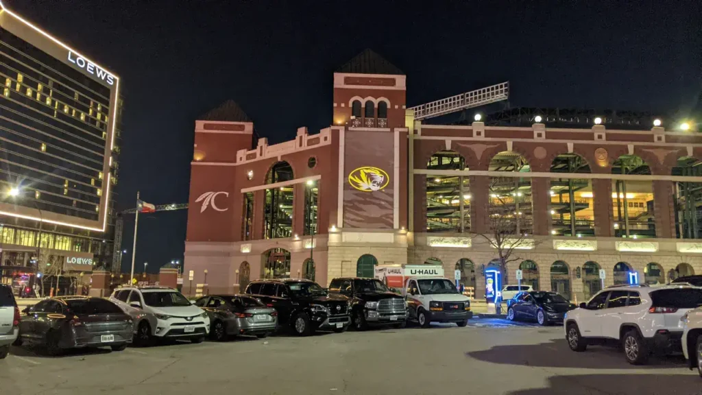 Nighttime view of a large brick building featuring a logo, surrounded by parked cars, with a modern hotel in the background, emphasizing urban advertising potential for guerrilla marketing strategies.