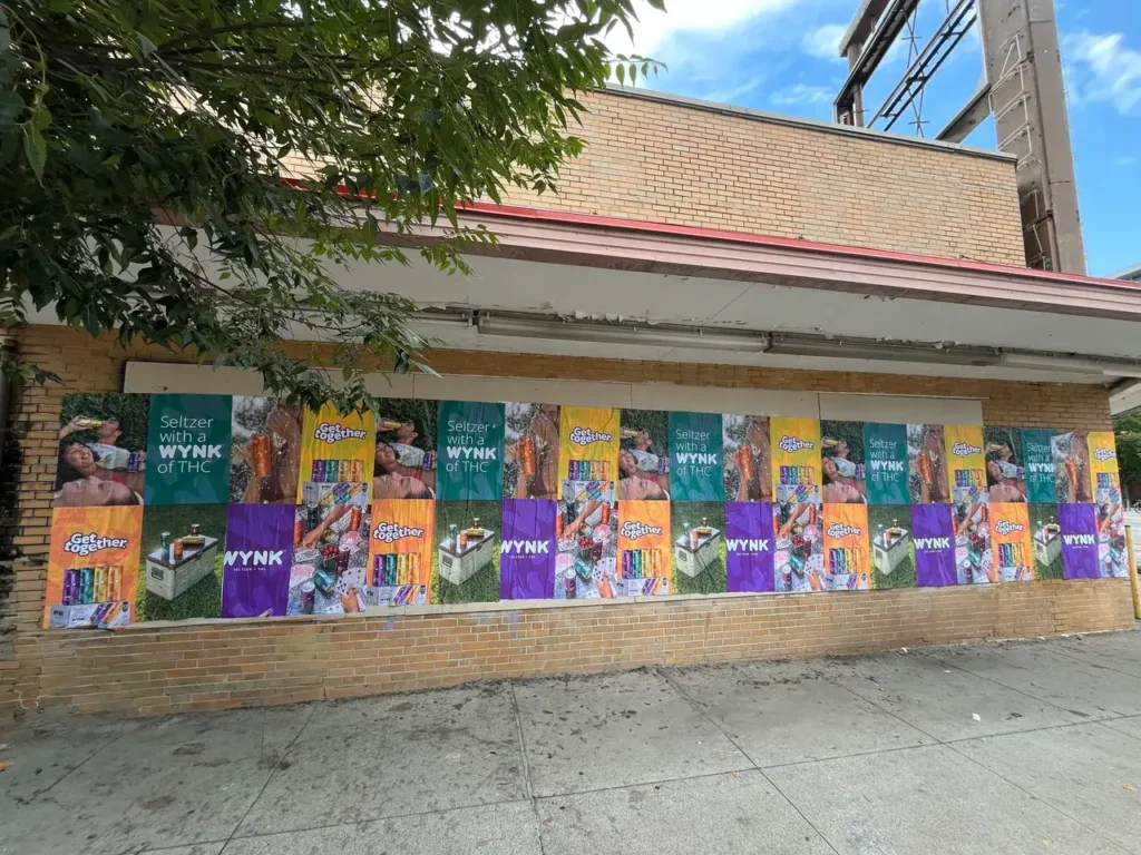 Colorful street advertising banners featuring "WYNK" seltzer with THC, displayed on a brick wall, promoting guerrilla marketing strategies in urban environments.