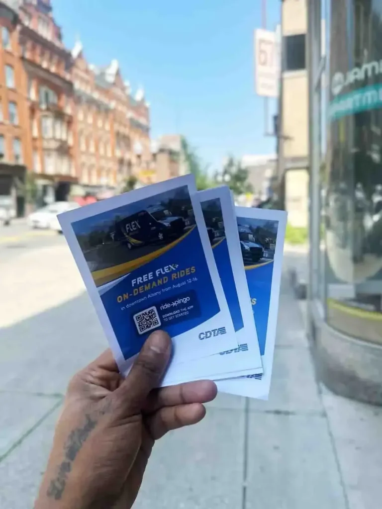 Hand holding promotional flyers for free on-demand rides, featuring a vehicle image and QR code, with a city street in the background.