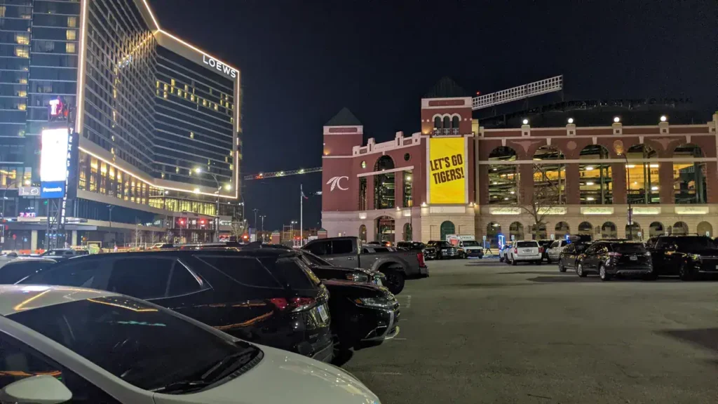 Nighttime view of a parking lot with parked cars, featuring a large building with illuminated signage for Loews and a prominent banner reading "LET'S GO TIGERS!" on a nearby stadium, highlighting the vibrant atmosphere of guerrilla marketing in urban settings.