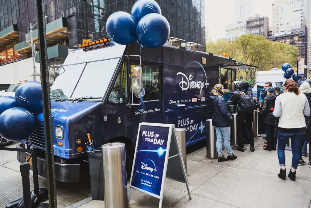 Disney+ food truck promoting streaming services, featuring blue balloons and a sign reading "PLUS UP YOUR DAY," with a crowd of people engaging nearby.