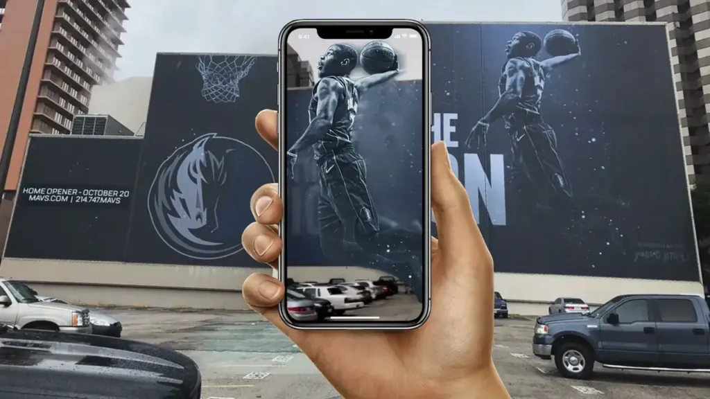 Hand holding smartphone displaying augmented reality basketball mural on building, showcasing interactive gaming experience for marketing.