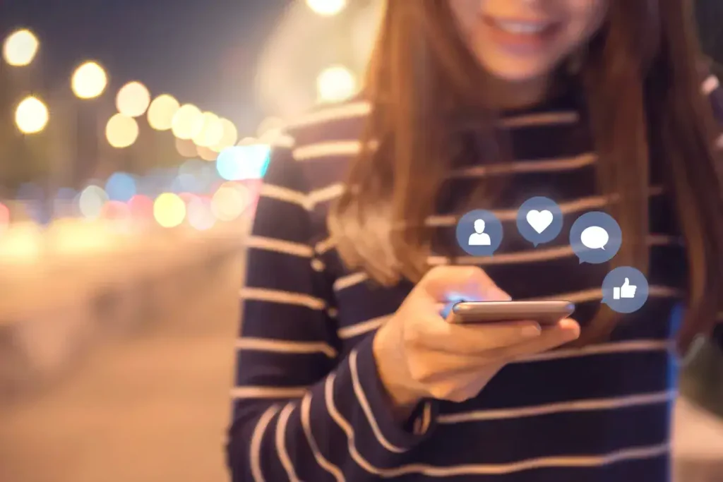 Woman using smartphone with social media icons, illuminated city background, representing digital marketing and audience engagement strategies.