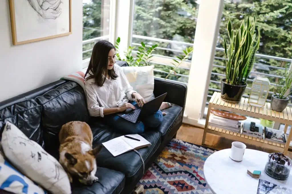 Woman working on laptop in cozy living room with dog, plants, and colorful decor, illustrating remote work and content creation.