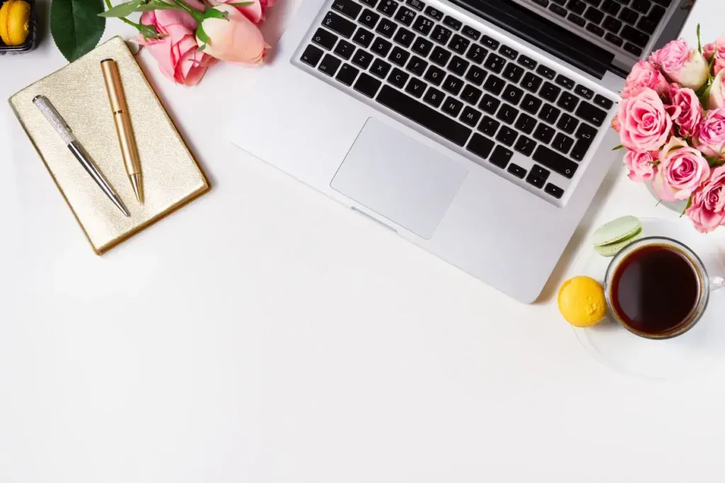 Laptop on a white desk surrounded by pink roses, a notebook, pens, a cup of coffee, and a lemon tart, symbolizing a creative workspace for marketing strategies.